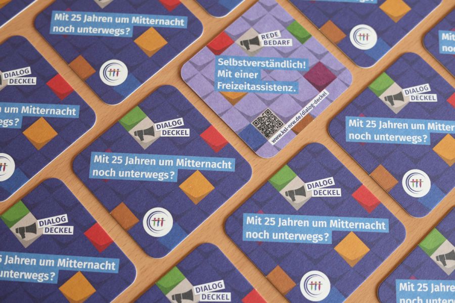 Mehrere Getränkeuntersetzer auf einem Tisch verteilt. Ein Deckel zeigt eine türkise Rückseite. Text auf der Vorderseite: Dialog-Deckel. Mit 25 Jahren um Mitternacht noch unterwegs? Text auf der Rückseite: Redebedarf. Selbstverständlich! Mit einer Freizeitassistenz.