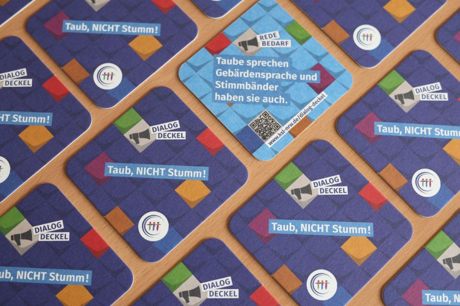 Mehrere Getränkeuntersetzer auf einem Tisch verteilt. Ein Deckel zeigt eine türkise Rückseite. Text auf der Vorderseite: Dialog-Deckel. Taub, nicht stumm! Text auf der Rückseite: Redebedarf. Taube sprechen Gebärdensprache und Stimmbänder haben sie auch. 
