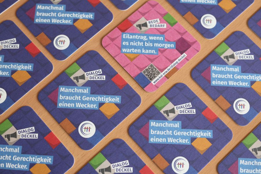 Mehrere Getränkeuntersetzer auf einem Tisch verteilt. Ein Deckel zeigt eine türkise Rückseite. Text auf der Vorderseite: Dialog-Deckel.  Manchmal braucht Gerechtigkeit einen Wecker. Text auf der Rückseite: Redebedarf. Eilantrag, wenn es nicht bis morgen warten kann.