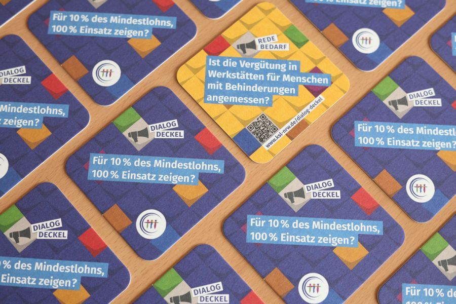Mehrere Getränkeuntersetzer auf einem Tisch verteilt. Ein Deckel zeigt eine türkise Rückseite. Text auf der Vorderseite: Dialog-Deckel. Für 10% des Mindestlohns, 100% Einsatz zeigen? Text auf der Rückseite: Redebedarf. Ist die Vergütung in Werkstätten für Menschen mit Behinderungen angemessen?