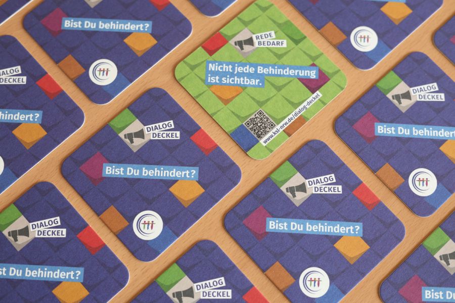 Mehrere Getränkeuntersetzer auf einem Tisch verteilt. Ein Deckel zeigt eine türkise Rückseite. Text auf der Vorderseite: Dialog-Deckel. Bist Du behindert? Text auf der Rückseite: Redebedarf. Nicht jede Behinderung ist sichtbar.