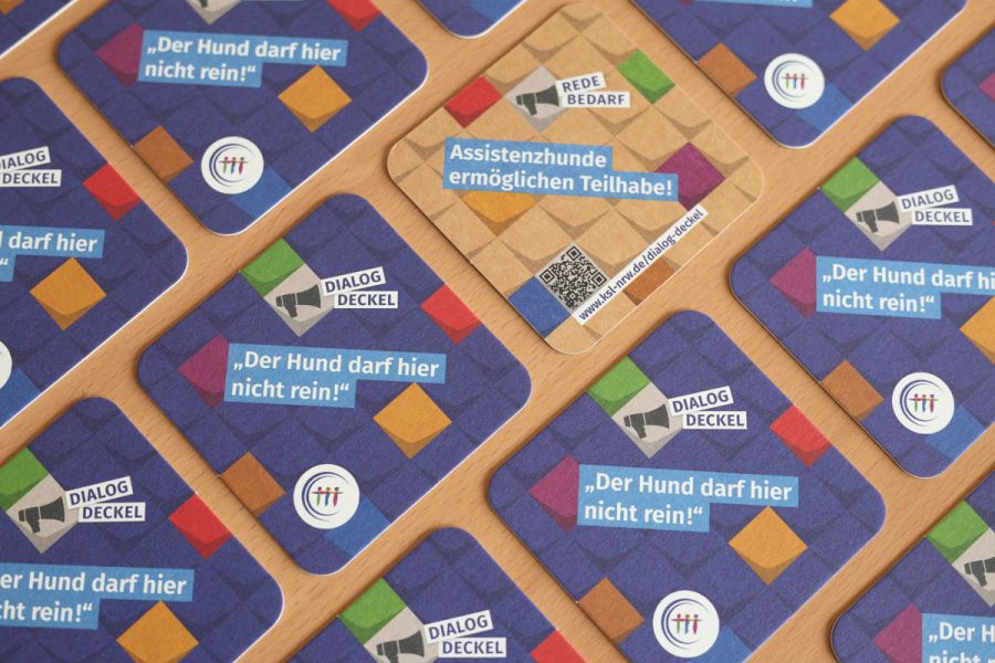 Mehrere Getränkeuntersetzer auf einem Tisch verteilt. Ein Deckel zeigt eine türkise Rückseite. Text auf der Vorderseite: Dialog-Deckel. „Der Hund darf hier nicht rein!“ Text auf der Rückseite: Redebedarf. Assistenzhunde ermöglichen Teilhabe!