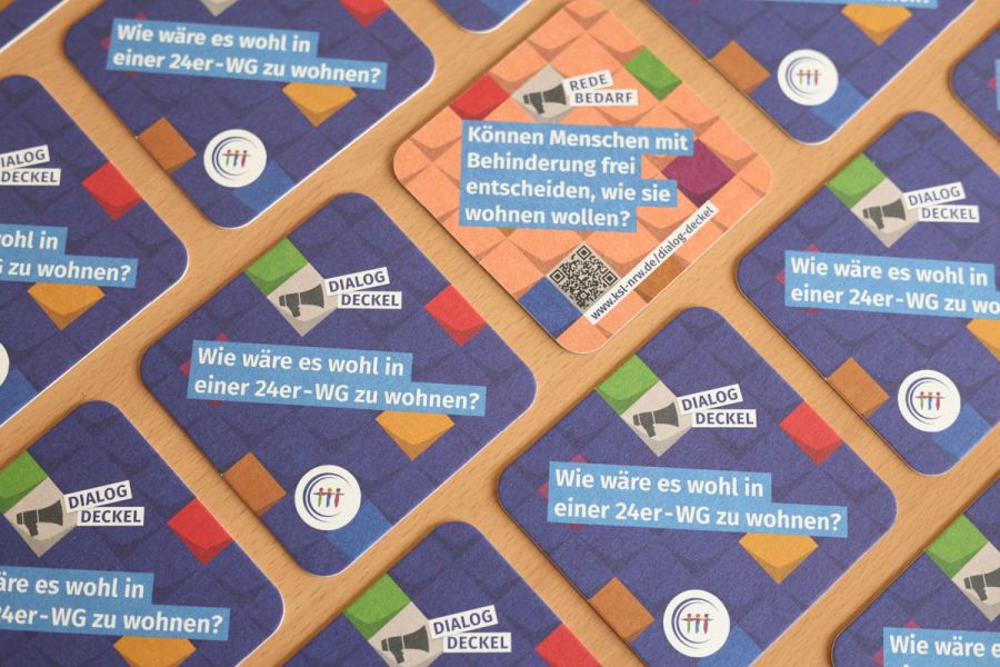 Mehrere Getränkeuntersetzer auf einem Tisch verteilt. Ein Deckel zeigt eine türkise Rückseite. Text auf der Vorderseite: Dialog-Deckel. Wie wäre es wohl in einer 24er-WG zu wohnen? Text auf der Rückseite: Redebedarf. Können Menschen mit Behinderung frei entscheiden, wie sie wohnen wollen?