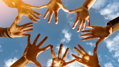 Acht Hände, die sich anfassen und einen Kreis bilden; im Hintergrund: blauer Himmel mit Wolken