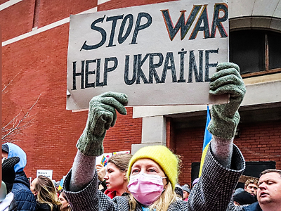 Eine Frau mit gelber Mütze zeigt ein Schild. Darauf steht Stop war, help Ukraine.