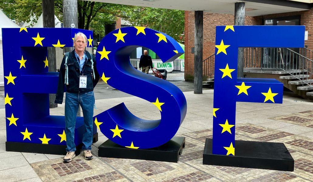 Andreas Tintrup, ein Mann mit längerem blonden Haar und Jeanskleidung, steht vor drei menschenhohen Buchstaben: ESF in blau mit gelben Sternen
