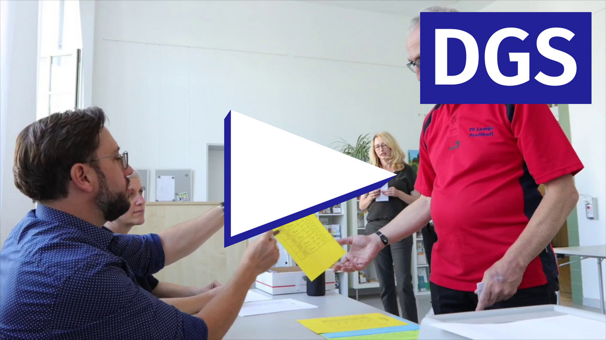 Eine Person bekommt einen Wahlzettel ausgehändigt, im Vordergrund ein Play-Knopf