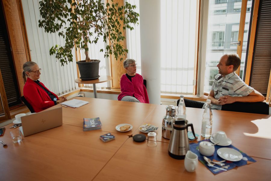 Zwei Frauen und ein Mann sitzen an einem Konferenztisch. Zu sehen sind Wibke Roth, eine Frau mit dunkelblondem Haar, Pferdeschwanz in rotem Blazer und Brille, eine Frau mit kurzen grauen Haar und Brille sowie pinker Strickjacke und ein Mann mit sehr kurzem Haar und gestreiftem kurzen Hemd 