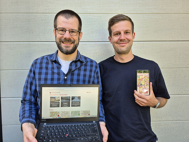 Foto von Gero Büskens (Junger dunkelhaariger Mann mit Bart links) und Niklas Büskens (junger dunkelhaariger Mann ohne ohne Bart) vor einer weißen Holzpanele.