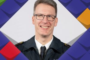 Ein Foto von Daniel Meyer, ein Manm mit Brille und kurzem dunkelblond-grauem Haar, der eine Feuerwehr-Uniform, ein weißes Hemd und eine dunkle Krawatte trägt. Er lächelt. Drumherum befinden sich Schmuckelemente der KSL.NRW