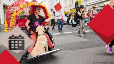 Eine Aufnahme von einer Mad Pride Parade aus den letzten Jahren. Eine Frau nimmt sitzend in einer Rikscha teil. Ihr Hund begleitet sie auf den Fußbrett.