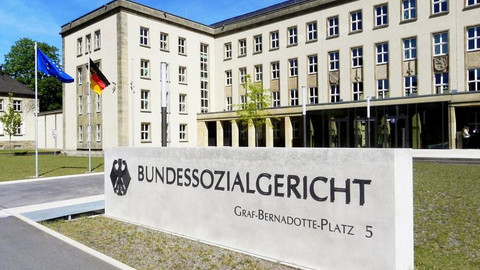 Das Foto zeigt den Eingangsbereich zum Bundessozialgericht. Eingrußes weißes Gebäude. Davor befindet sich eine weiße Tafel mit der Aufschrift Bundessozialgericht.