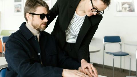 In einem Büro: Eine Frau zeigt einem offenbar blinden Mann etwas in einem aufgeschlagenen Buch. Dazu führt sie seine Hand. 