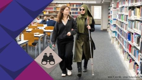 Kübranur Binek, eine Frau mit Brille und Langstock hakt sich bei ihrer Assistenz Madita Laumen unter. Madita Laumens Blick ist suchend auf das Bücherregal im rechtswissenschaftlichen Seminar gerichtet.